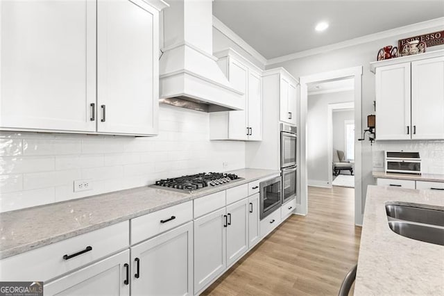 kitchen featuring white cabinetry, light stone counters, custom range hood, appliances with stainless steel finishes, and crown molding