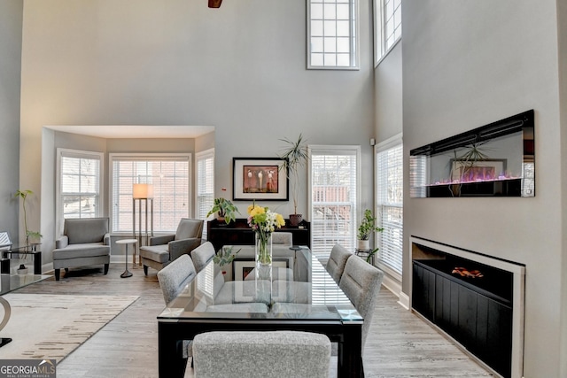 dining space featuring a warm lit fireplace, baseboards, light wood-style flooring, and a high ceiling