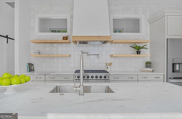 kitchen featuring stainless steel fridge with ice dispenser, premium range hood, a barn door, light stone counters, and open shelves