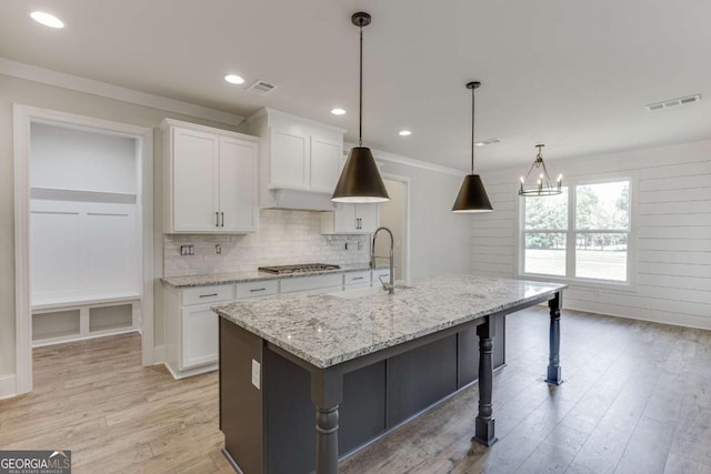 kitchen with light wood-type flooring, a center island with sink, hanging light fixtures, light stone countertops, and white cabinets