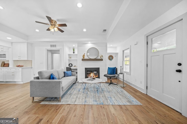 living room featuring light hardwood / wood-style flooring, built in shelves, a tray ceiling, a large fireplace, and ceiling fan