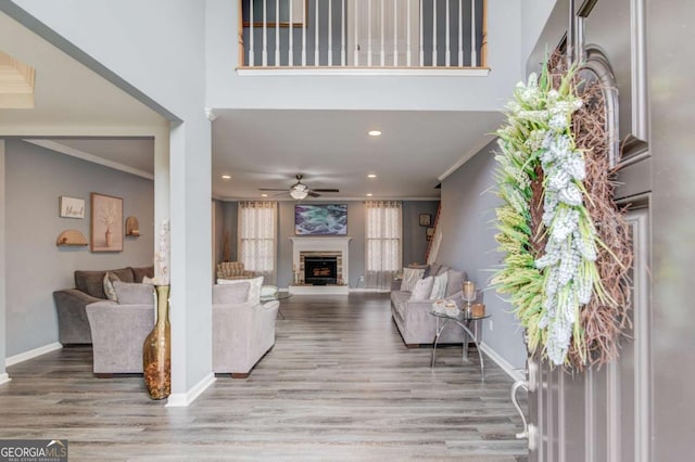 living room featuring a fireplace with raised hearth, crown molding, baseboards, wood finished floors, and a ceiling fan