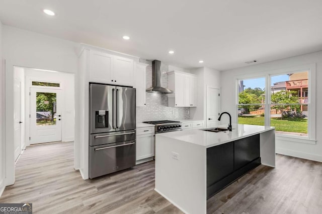 kitchen featuring high end appliances, an island with sink, wall chimney range hood, sink, and white cabinetry