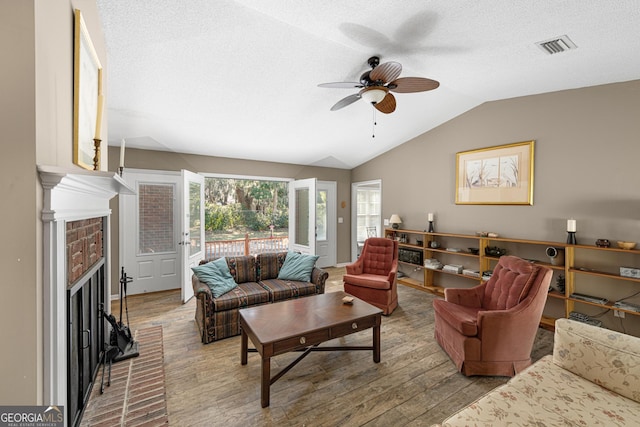 living room with a textured ceiling, ceiling fan, light hardwood / wood-style floors, vaulted ceiling, and a brick fireplace