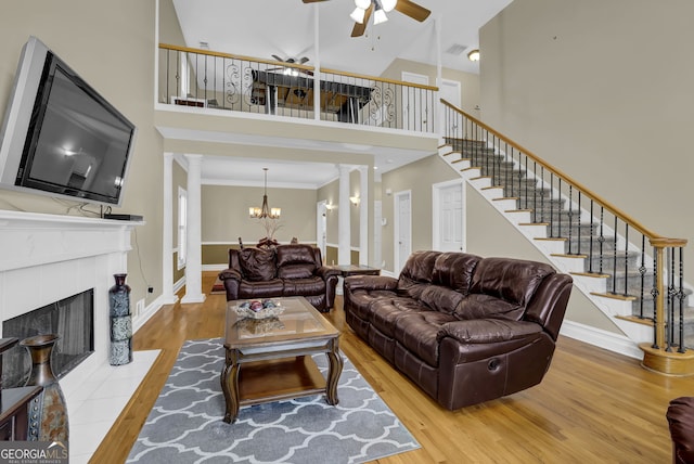 living area with wood finished floors, stairs, ornate columns, a fireplace, and ceiling fan with notable chandelier