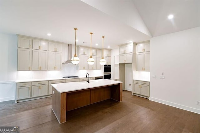 kitchen with gas cooktop, hanging light fixtures, double wall oven, wall chimney range hood, and a kitchen island with sink