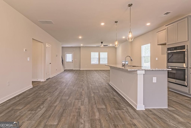 kitchen with double oven, gray cabinetry, light stone countertops, an island with sink, and dark wood finished floors