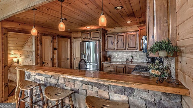 kitchen featuring a breakfast bar, stainless steel refrigerator with ice dispenser, wooden ceiling, hanging light fixtures, and wooden walls