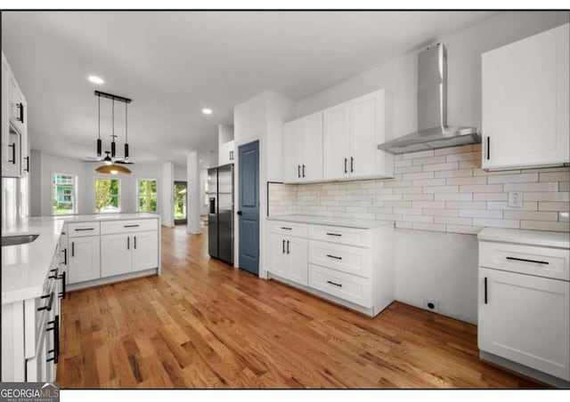 kitchen featuring wall chimney range hood, white cabinets, hanging light fixtures, and stainless steel refrigerator with ice dispenser