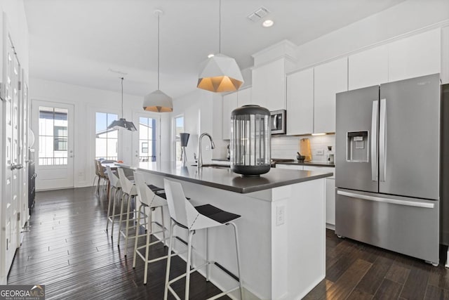 kitchen featuring stainless steel appliances, dark countertops, a center island with sink, and white cabinetry