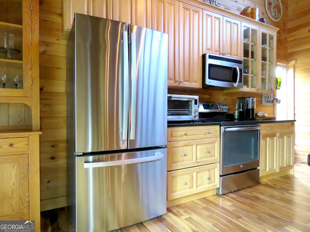 kitchen featuring light wood finished floors, wooden walls, dark countertops, glass insert cabinets, and appliances with stainless steel finishes