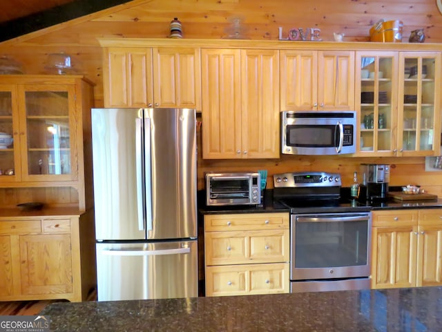 kitchen featuring a toaster, stainless steel appliances, wood walls, dark stone counters, and glass insert cabinets