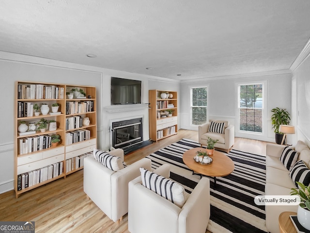 living room with built in shelves, a glass covered fireplace, crown molding, and light wood finished floors