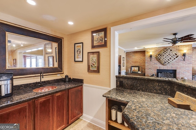 interior space with a brick fireplace, wainscoting, a sink, ceiling fan, and dark stone countertops