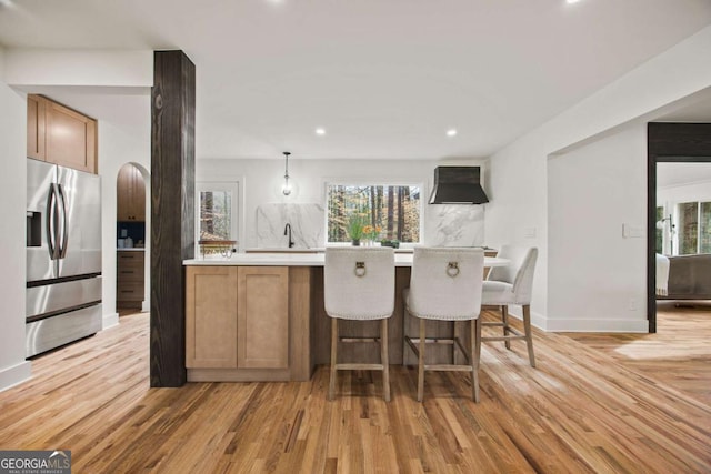 kitchen with light wood-type flooring, kitchen peninsula, stainless steel fridge, wall chimney exhaust hood, and pendant lighting