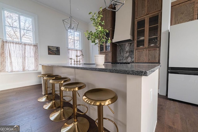 kitchen featuring dark wood-type flooring, custom range hood, refrigerator, decorative backsplash, and a breakfast bar