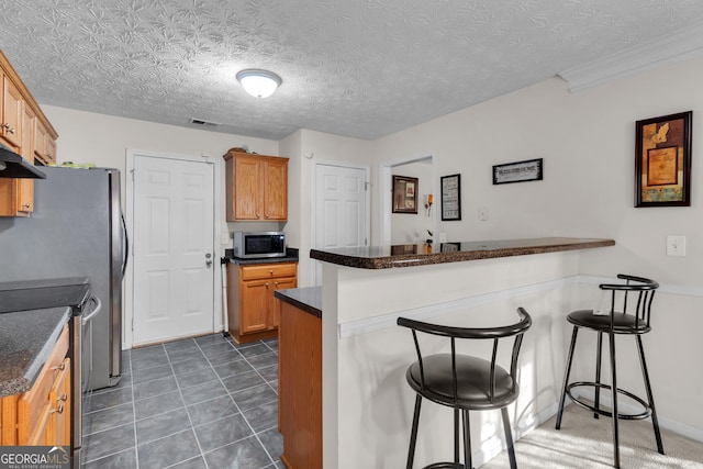 kitchen featuring under cabinet range hood, a peninsula, stainless steel appliances, dark tile patterned floors, and a kitchen bar