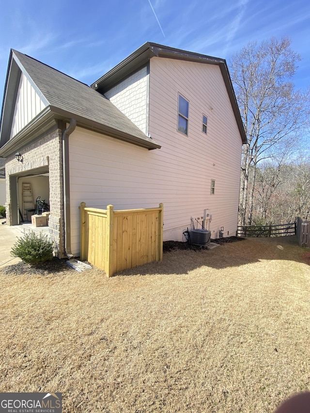 view of side of home featuring fence, an attached garage, board and batten siding, central air condition unit, and brick siding