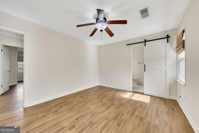 unfurnished bedroom featuring ceiling fan, a barn door, light wood-type flooring, and ensuite bath