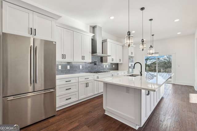 kitchen featuring appliances with stainless steel finishes, white cabinetry, wall chimney range hood, hanging light fixtures, and an island with sink