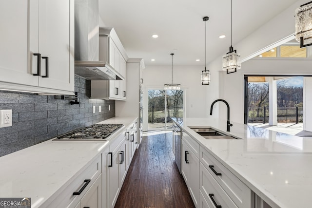 kitchen with hanging light fixtures, light stone countertops, white cabinets, wall chimney range hood, and sink