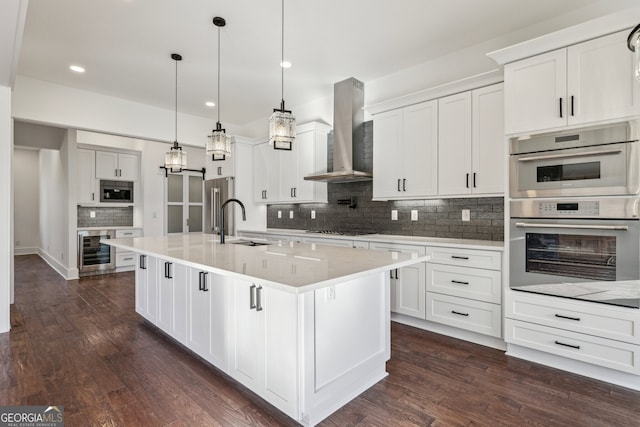 kitchen featuring beverage cooler, decorative light fixtures, wall chimney range hood, stainless steel appliances, and an island with sink