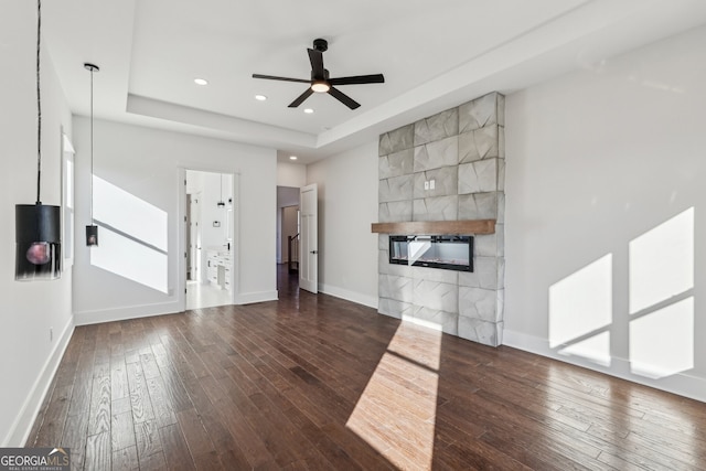 unfurnished living room featuring a tile fireplace, ceiling fan, a tray ceiling, and dark hardwood / wood-style flooring