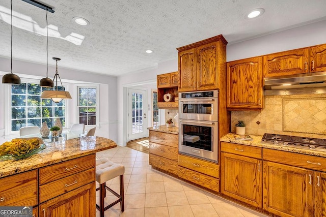 kitchen with light tile patterned floors, under cabinet range hood, stainless steel appliances, brown cabinets, and tasteful backsplash