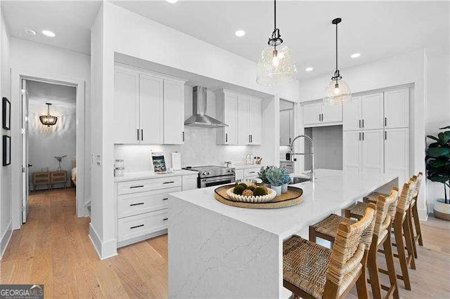 kitchen with a center island with sink, stainless steel electric range oven, white cabinetry, wall chimney exhaust hood, and hanging light fixtures