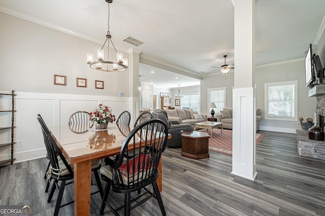 dining room featuring visible vents, dark wood-type flooring, crown molding, a stone fireplace, and ceiling fan with notable chandelier