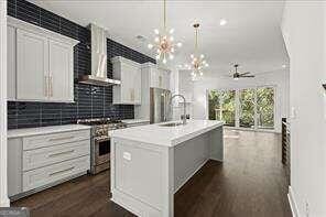 kitchen with a center island with sink, hanging light fixtures, high end stainless steel range oven, white cabinets, and wall chimney range hood