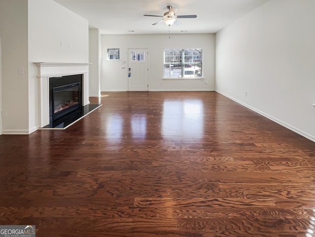 unfurnished living room featuring dark wood-type flooring, a glass covered fireplace, ceiling fan, and baseboards