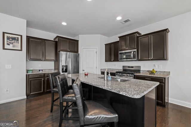 kitchen featuring dark brown cabinetry, stainless steel appliances, dark wood-style flooring, a sink, and light stone countertops