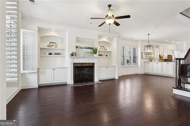 unfurnished living room with a premium fireplace, built in shelves, ceiling fan with notable chandelier, and dark wood-type flooring