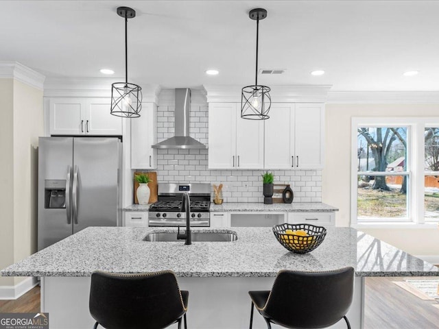 kitchen featuring a center island with sink, wall chimney exhaust hood, stainless steel appliances, decorative light fixtures, and white cabinetry