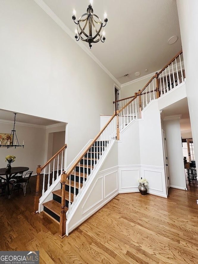 stairs with a wainscoted wall, crown molding, a towering ceiling, wood finished floors, and a chandelier