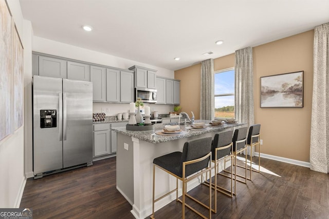 kitchen featuring appliances with stainless steel finishes, a center island with sink, gray cabinetry, a breakfast bar area, and dark hardwood / wood-style flooring