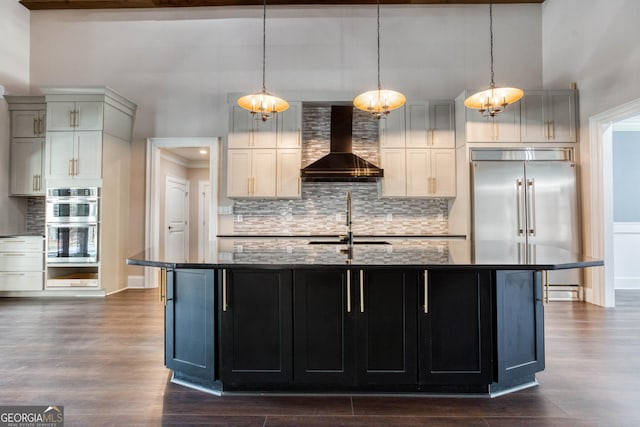 kitchen with an island with sink, dark wood-style floors, appliances with stainless steel finishes, wall chimney range hood, and dark stone counters