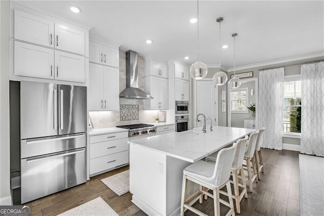 kitchen with pendant lighting, stainless steel appliances, white cabinetry, an island with sink, and wall chimney exhaust hood