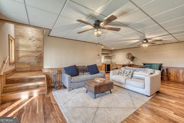 living area with a drop ceiling, wainscoting, ceiling fan, stairway, and light wood-style floors