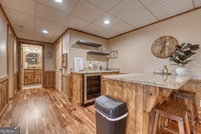 bar featuring wine cooler, a drop ceiling, bar area, light wood-style floors, and wainscoting