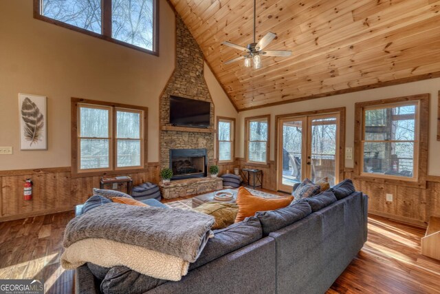 living room featuring a stone fireplace, a wainscoted wall, ceiling fan with notable chandelier, wood finished floors, and wood ceiling