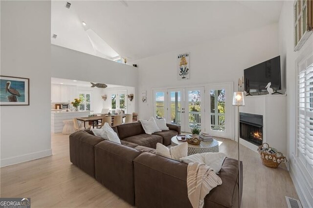 living area featuring high vaulted ceiling, light wood-type flooring, a warm lit fireplace, and visible vents