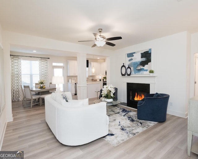 living room featuring light wood-type flooring, a warm lit fireplace, ceiling fan, and baseboards