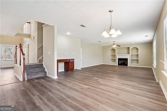 unfurnished living room with built in features, wood finished floors, stairway, baseboards, and ceiling fan with notable chandelier