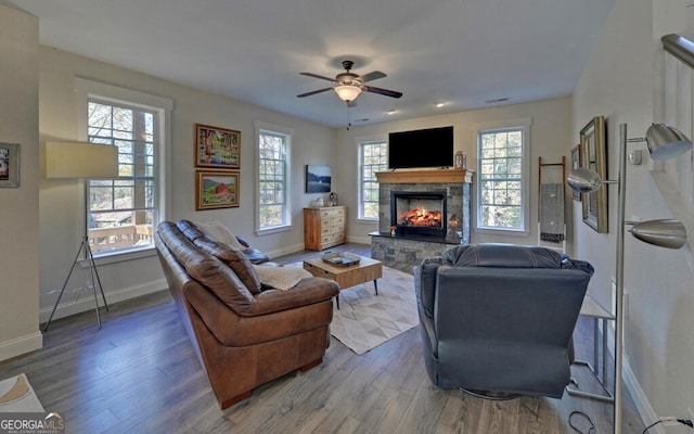 living area featuring ceiling fan, a stone fireplace, dark wood finished floors, and baseboards