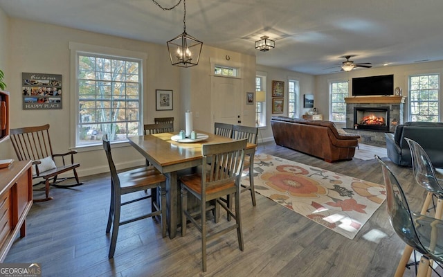 dining space featuring ceiling fan with notable chandelier, a stone fireplace, wood finished floors, and baseboards