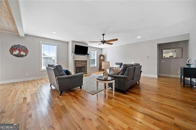 living room featuring baseboards, light wood-style flooring, ceiling fan, a stone fireplace, and beam ceiling