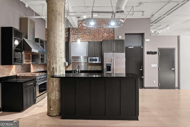 kitchen featuring dark countertops, glass insert cabinets, stainless steel appliances, wall chimney range hood, and a sink