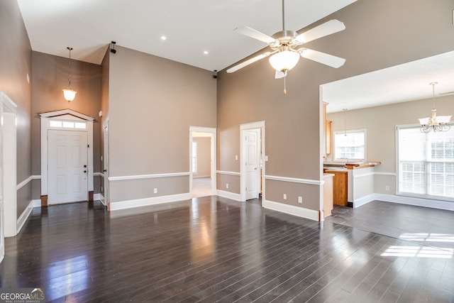 unfurnished living room featuring dark wood-style flooring, a towering ceiling, baseboards, and ceiling fan with notable chandelier